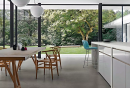Salon z dużymi oknami z widokiem na las, białą wyspą kuchenną, drewnianym stołem i krzesłami oraz podłogą imitującą beton Florim Creative Design Studios Concreate
