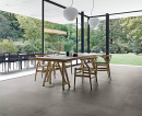 Salon z dużymi oknami z widokiem na las i taras ogrodowy, drewnianym stołem i krzesłami oraz podłogą imitującą beton Florim Creative Design Studios Concreate