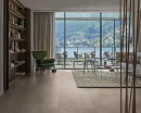 Salon z dużymi oknami z widokiem na balkon, zasłonami, drewnianym regałem, zielonym fotelem, jasną kanapą i płytkami podłogowymi imitującymi beton Florim Creative Design Studios Sand