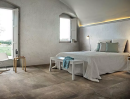 Aranżacja sypialni z dużym łóżkiem, szarymi ścianami przypominającymi beton oraz płytkami podłogowymi imitującymi kamień Florim Luxury Design Matières Brun