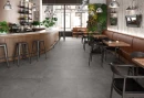 Wnętrze restauracji z drewnianą ladą barową, stolikami, czarnymi krzesłami i płytkami podłogowymi imitującymi beton Colorker Boxer & Uniq Graphite