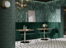 Wnętrze nowoczesnej restauracji z drewnianymi stolikami, zielonymi krzesłami, kanapą z panelami ściennymi tapicerowanymi oraz płytkami w kolorze zielonym Paradyż Ceramika Monpelli Emerald Mix oraz mozaiką Paradyż Monpelli Emerald Murano
