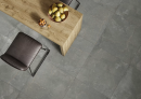 Wnętrze z drewnianą wyspą, krzesełkami oraz płytkami podłogowymi, w kolorze grafitowym, imitującymi kamień Paradyż Ceramika Fillstone Graphite