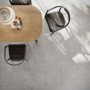Wnętrze z drewnianym stołem, czarnymi krzesłami i płytkami podłogowa imitująca kamień, w kolorze szarym Flaviker Double Plain Grey Naturale