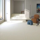 Podłoga salonu oraz schody wyłożone płytkami Arcana Spuma-R Panna 80x80. Duże okno na ścianie rozjaśnia całe przestronne pomieszczenia a kolorowe dekoracje w postaci brązowego fotela i niebieskiego podnóżka uzupełniają całą kompozycję