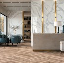 Recepcja hotelu z drewnianą podłogą ułożoną w jodełkę, fotelami wypoczynkowymi, brązową kamienną ladą oraz płytkami na ścianie imitującymi marmur w kolorze białym z szarą żyłą Mirage Jewels Calacatta Reale Lucido JW02