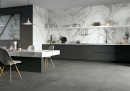 Kuchnia w stylu glamour połączona z jadalnią z długim marmurowym stołem, białymi krzesłami, grafitowymi szafkami, szarą podłogą oraz płytkami na ścianie imitującymi marmur Mirage Jewels Statuario Lunensis Lucido JW12