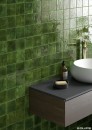 Hiszpańska zielona płytka ceramiczna w połysku Equipa Menorca Tourmaline 10x10 na ścianie łazienki. Na płytkach zamontowana szafka umywalkowa w ciemnym brązowym kolorze. Biała umywalko oraz złota bateria umywalkowa