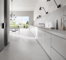 Kuchnia połączona z jadalnią, z jasnymi szafkami, białym stołem, dużym oknem z widokiem na balkon i płytkami podłogowymi imitującymi kamień Leonardo Ceramica Ashima ASHM 60W RM