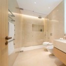 Łazienka wyłożona kremowymi płytkami marmurowymi Marazzi Allmarble Crema Marfil Natural MELE 60x120 cm, z dwoma białymi sedesami, szklanym parawanem prysznicowym oraz chromowym wieszakiem na ręczniki, na którym znajduję się biały ręcznik