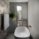 Łazienka z dużą owalną białą wanną, nad którą widnieje złota bateria, wyłożona matowymi płytkami ściennymi imitującymi beton, Marazzi Cementum Wall Nickel w prawej części oraz z lewej patchworkowymi płytkami