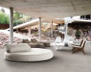 Taras wyłożony strukturalnymi szarymi płytkami tarasowymi imitującymi beton, Marazzi Cementum20 Nickel Structured Surface, z białą dużą okrągłą kanapą, małym białym stolikiem, dwoma materiałowymi leżakami oraz czterema krzesłami