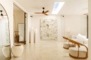 Przestronny salon z drewnianymi fotelami, białą sofą, owalnym dużym lustrem w rogu, wyłożony połyskujacymi kremowymi płytkami imitującymi marmur Marrazi Grande Marble Look Crema Marfil Lux
