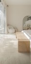 Przestronna sypialnia z marmurową podłogą, beżowym stolikiem wykonanym z płytek Marazzi Grande Marble Look Onice Beige Lux i białym fotelem oraz dużym owalnym lustrem w rogu