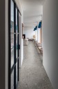 Nowoczesny korytarz z szarą podłogą z płytek Marazzi Mystone Ceppo Di Gre Grey Natural, oświetlony naturalnym światłem z przeszklonych drzwi po lewej stronie, wzdłuż prawej ściany ciągnie się drewniana ławka z poduszkami i niebieskimi lampami