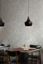 Detal ściany w jadalni, pokrytej szarą mozaiką ceramiczną Marazzi Naturalia Grigio Mosaico, składającą się z wielu sześciokątnych elementów, przed ścianą stoi stół z czarnymi krzesłami, herbatą i talerzem z jedzeniem