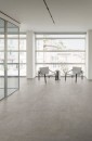 Minimalistycze wnętrze z widokiem na miasto z podłogą wyłożoną płytkami Marazzi Plaza Multicolor o zmiennych odcieniach beżu i szarości doskonale imituje beton, na tle panoramicznych okien ustawiono dwa nowoczesne fotele w jasnym kolorze