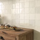 Drewniana komoda w kuchni na której leży deska z jedzeniem oraz miskami, za komodą na ścianie położone są płytki Vives Root Artis Pearl 13x13 w odcieniu kremowym