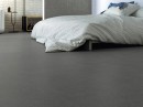Sypialnia z płytką gresową, podłogową imitującą beton Cifre Acron Anthracite Mate N-Plus SI Rect. 90x90, z niskim łóżkiem nakrytym jasną pościelą w kratkę oraz ozdobnymi poduszkami, z minimalistyczną konsolą.