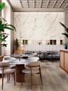 Wnętrze eleganckiej restauracji z drewnianymi stolikami, marmurową dekoracyjną ścianą oraz płytkami podłogowymi drewnopodobnymi Porcelanosa Bruselas Walnut