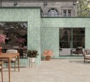 Gresowa dekoracyjna płytka ścienna w zielonym kolorze Gayafores Deco Artis Salvia 32x62,5 na elewacji budynku. na pierwszym planie stoliki, krzesła oraz kwity doniczkowe na tarasie.