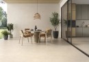 Płytka gresowa, podłogowa w kolorze beżowym, wykończenie powierzchni matowe, Geotiles Talo Arena Mate Rect. 60x120 położona na podłodze tarasowej, gdzie znajduje się stół z czterem krzesłami, nad nimi lampa oraz przeszklone wejście do domu