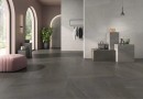 Płytka gresowa, podłogwa w kolorze grafitowym imitująca kamień, wykończenie powierzchni matowe, Geotiles Eddystone Gris Mate 60,8x60,8 położona w nowoczesnym salonie, w którym znajduje sie różowa sofa, wieszaki na ubrania oraz okna z widokiem na ogród