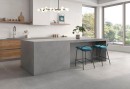 Płytka ścienna w kolorze szarym, wykończenie powierzchni matowe, Geotiles Portland Perla 36x80 położona na ścianie w kuchni gdzie znajduje się zlewozmywak, szafki kuchenne w brązowym kolorze, dwa krzesła oraz duże okno z widokiem na park