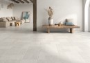 Płytka gresowa, podłogowa w kolorze szarym, wykończenie powierzchni satynowe, Geotiles Montpellier Perla Satyna Rect. 60x120 położona na ścianie w pokoju gdzie znajduje się wypoczynek w jasnym kolorze, lampa wisząca oraz stolik na kwiaty, świeczki