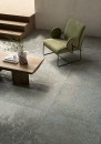 Ciemnoszara gresowa płytka podłogowa imitująca betonowe lastryko Fioranese Alchimia Grigio Azzurro Naturale 90,6x90,6 AL907R na podłodze salonu. Drewniany stolika kawowy w jasno brązowym kolorze oraz zielony welurowy fotel na metalowych czarnych nogach