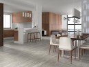 Wnętrze kuchni połączonej z jadalnią z drewnianymi frontami szafek, drewnianym stołem, jasnymi krzesłami i szarymi płytkami podłogowymi drewnopodobnymi Porcelanosa Smart Tanzania Silver