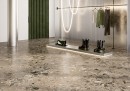 Gresowa podłogowa szara płytka imitująca lastryko Fioranese I Ciottoli Sassi Antracite Naturale 90,6x90,6 LMS900R na podłodze salonu mody w połączeniu z zielonymi elementami oraz czarnymi wieszakami