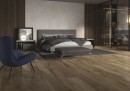 Płytka gresowa, podłogowa imitująca drewno w kolorze brązowym, wykończenie powierzchni matowe, Cerrad Guardian Wood Brown 19,3x120,2 położona w sypialni gdzie znajduje się fotel, duże łóżko ora przeszklona garderoba