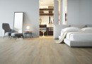 Płytka gresowa, podłogowa imitująca drewno w kolorze brązowym, wykończenie powierzchni matowe, Cerrad Mattina Beige Rect. 29,7x120,2 położona na podłodze w salonie gdzie znajduje się fotel, lustro oparte o ścianę, duże łóżko oraz wejście do garderoby