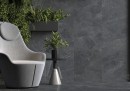 Płytka gresowa, podłogowa immitująca kamień w kolorze grafitowym, wykończenie powierzchni satynowe, Cerrad Distinct Graphite Silky Cristal 59,7x119,7 położona na podłodze oraz ścianie w pomieszczeniu gdzie znajduje się jasny fotel a za nim ozdobne rośliny