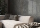 Płytka gresowa, podłogowa imitująca beton w kolorze grafitowym, wykończenie powierzchni satynowe, Cerrad Textural Anthracite Silky Cristal Rect. 59,7x59,7 położona na ścianie w pokoju gdzie znajduje się jasna kanapa oraz roślina ozdobna