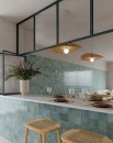 Płytka ścienna, dekoracyjna w kolorze zielonym, wykończenie powierzchni w połysku, Gayafores Deco Amaris Green 32x62,5 położona na ścianie w kuchni gdzie znajdują się krzesła, blat jasna kanapa oraz wiszące lampy