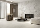Gresowa podłogowa płytka imitująca beton Fioranese The Blend Concrete Ivory Naturale 120,8x120,8 GC121R na podłodze jasnego, przestronnego salonu w zestawieniu z czarnym stolikiem, jasnymi meblami, kontrastującym czarnym kominkiem oraz kamienną ścianą