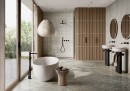Gresowa podłogowa szara płytka imitująca beton Fioranese The Blend Concrete Silver Levigato-Matt 60,4x60,4 GC603LR na ścianie nowoczesnej łazienki w połąćzeniu z drewnianymi elementami dekoracyjnymi, czarną armaturą podtynkową oraz wolnostojącą wanną