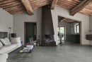 Przestronny salon w stylu rustykalnym z wysokim, drewnianym sufitem z belkami, gdzie podłoga wyłożona jest matowymi, szarymi płytkami antypoślizgowymi Marazzi Mystone Pietra Ligure Cenere R10, imitującymi kamień, centralnym punktem wnętrza jest kominek
