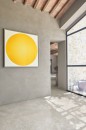 Jasne wnętrze utrzymane w stylu śródziemnomorskim, z podłogą wyłożoną prostokątnymi, jasnymi płytkami ceramicznymi Marazzi Mystone Pietra Ligure Bianco 60x120 cm, na strukturalnej, beżowo-szarej ścianie wisi duży, nowoczesny obraz z żółtym kołem