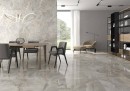 Płytka gresowa, podłogowa imitująca kamień w kolorze beżowym, wykończenie powierzchni w połysku, Pamesa Lux Noor Almond Polished 60x120 położona na podłodze w salonie gdzie znajduje się stół jadalny i krzesła oraz fotel przy oknie z widokiem na ogródek