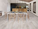Gresowa podłogowa płytka imitująca białe drewnoSaime Alpi Bianco Naturale 30x120 8600328 na podłodze otwartej i przestronnej jadalni w otoczeniu dużego stołu jadalnego, drewnianych krzeseł oraz nowoczesnej kuchni w drewnianym wykończeniu