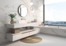 Płytka ścienna imitująca marmur w białym kolorze, wykończenie powierzchni satynowe struktualne, Pamesa Rlv Calacata Gold satin 60x120 położona na ścianie w łazience gdzie znajduje się wanna przy dużym oknie oraz półka z umywalką i wiszące lustro
