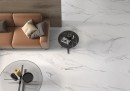 Płytka gresowa, podłogowa imitująca marmur w białym kolorze, wykończenie powierzchni w połysku, Pamesa Calacata White Polished 120x120 położona na podłodze w salonie gdzie znajduje się brązowa kanapa, szklany stolik do kawy oraz drewniana ściana