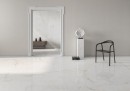 Płytka gresowa, podłogowa imitująca kamień w białym kolorze, wykończenie powierzchni w połysku, Pamesa Crystal White Polished 120x120 położona na podłodze w pomieszczeniu gdzie znajduje się czarne krzesło, posąg oraz przejście do drugiego pokoju