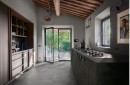Nowoczesna kuchnia w rustykalnym stylu, z podłogą wyłożoną szarymi płytkami gresowymi Marazzi Mystone Pietra Ligure Cenere 60x120 MPK6, imitującymi kamień, wnętrze z drewnianymi belkami sufitowymi, ciemnymi szafkami kuchennymi, zintegrowaną płytą gazową