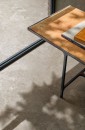 Bliskie ujęcie podłogi wyłożonej beżowymi płytkami gresowymi Marazzi Mystone Pietra Ligure Beige 60x60 cm MPK8, imitującymi kamień w ciepłym odcieniu, na podłogę pada cień liści i naturalne światło, a w prawym górnym rogu widoczny jest narożnik stolika