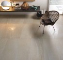 Gresowa beżowa płytka podłogowa imitująca kamień Saime Artica Beige Naturale 60x120 8600628 na podłodze salonu. Czarna wisząca półka z dekoracjami i lampką. Wiklinowy fotel z metalowymi nogami.