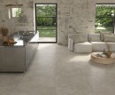 Gresowa podłogowa płytka imitująca beton Saime Avalon Gris Naturale Modulo 8690024 na podłodze salonu z aneksem kuchennym. Stara ceglana ściana, duże okna i szklane drzwi w czarnej ramie. Jasna, beżowa kanapa, okrągły drewniany stolik kawowy.